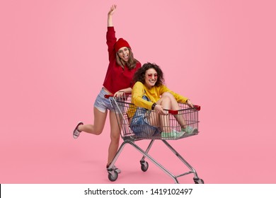 Two cheerful young women in colorful trendy outfits smiling and having fun with trolley during shopping against pink background - Shutterstock ID 1419102497
