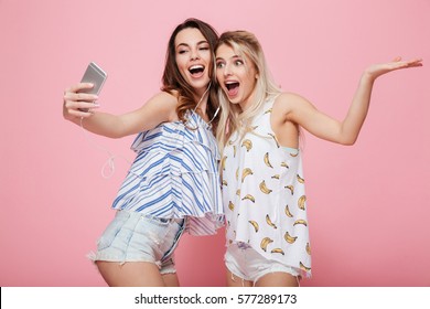 Two cheerful young women with earphones listening to music and talking selfie over pink background