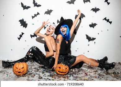 Two cheerful young women in leather halloween costumes posing with curved pumpkins over bats and confetti background