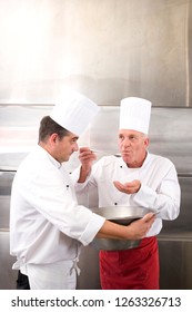 Two Chefs Wearing Whites In Commercial Kitchen Tasting Food