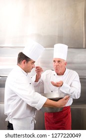 Two Chefs Wearing Whites In Commercial Kitchen Tasting Food