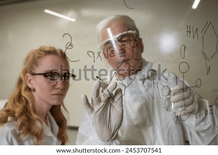 Two chemist colleagues write formulas on glass. Caucasian elderly man and young woman brainstorming.