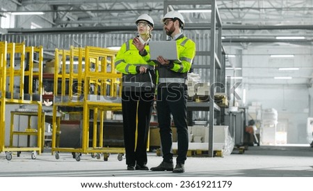 Two chief engineers are discussing project and looking at laptop. Man and woman in yellow vests communicate at construction site. Employees gesturing on background of racks.
