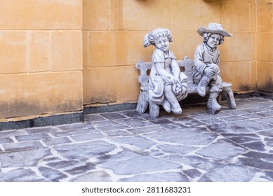Two Child Statues Sitting on a Bench Against a Rustic Wall in a Sunny Courtyard
