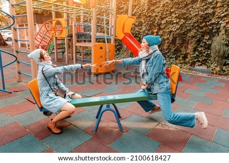 Two childish happy woman friends or teenage girl having fun on seesaw on a playground. Concept of psychology of the new generation and relations