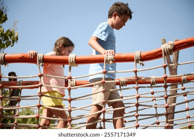 Two children carefully walk across a rope bridge in an outdoor playground. The scene is bright and emphasizes a sense of adventure and playfulness under a clear sky. - Powered by Shutterstock