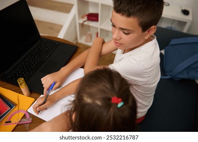 Two children collaborate on school work with a laptop on the desk. They are focused on writing and learning in a cozy study environment. - Powered by Shutterstock