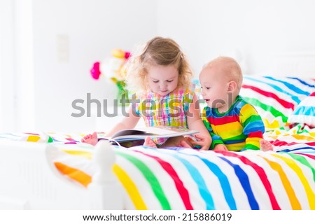 Two children, curly little toddler girl and a funny baby boy, brother and sister, reading a book in a sunny bedroom on a wooden white bed with colorful rainbow bedding enjoying a nice weekend morning