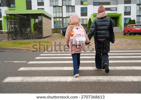 Two children dressed in winter jackets and backpacks walk hand in hand across a marked crosswalk outside a modern school building. The urban setting features parked cars, bicycles, and an overcast.