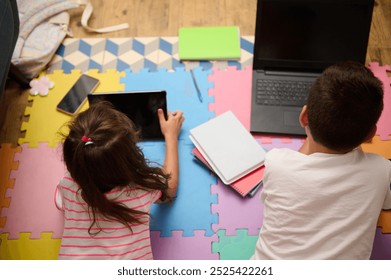 Two children engage with digital devices, lying on colorful foam mats. A casual learning and play setting indoors is depicted, emphasizing technology use. - Powered by Shutterstock