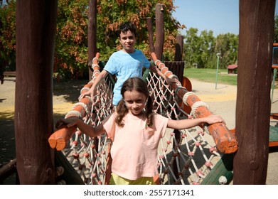 Two children enjoy a sunny day playing on a rope bridge at the park. Laughter and adventure fill the air as they explore the playground together. - Powered by Shutterstock