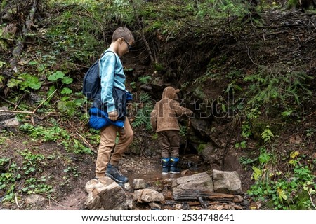 Two children exploring forest path, one stepping through small stream, both dressed in warm clothes and backpacks. Childhood adventure, nature exploration, and outdoor activities for kids in autumn.