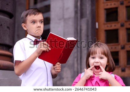 Two children having fun in front of school doors. Boy with jokingly serious face reading from textbook, girl makes funny face grimace. Focus on boy's face. Friends enjoying reunion on first shool day.