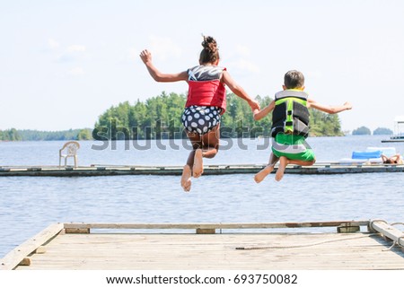 Two children are holding hands, jumping off a wooden dock into the lake water. They are wearing life jackets. There is one girl and one boy holding hands as they plunge into the water. 