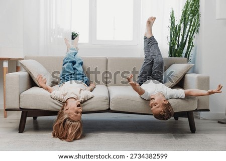 Two children laying upside down on a beige sofa in a bright living room with large windows and plants. They laugh and play, expressing joy and carefree childhood moments indoors.