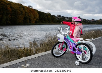 Two children on a lakeside pathway pause during their bike ride to admire the view. They wear colorful helmets and jackets, indicating a joyous outdoor adventure amid autumn foliage. - Powered by Shutterstock