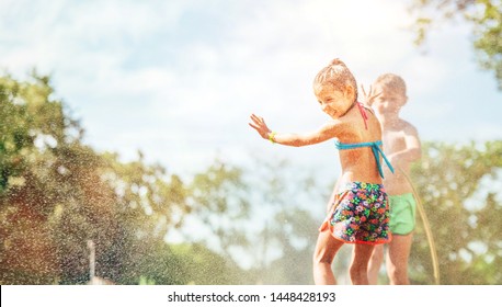 Two Children Play With Sprinkling Water In Summer Garden