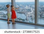 Two children point at landmarks while looking out a large window overlooking a cityscape, London, UK