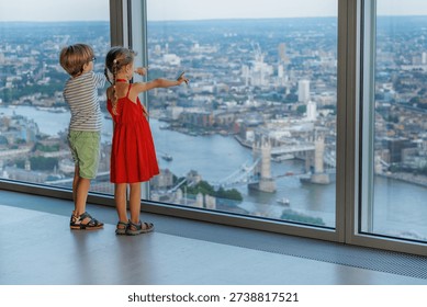 Two children point at landmarks while looking out a large window overlooking a cityscape, London, UK