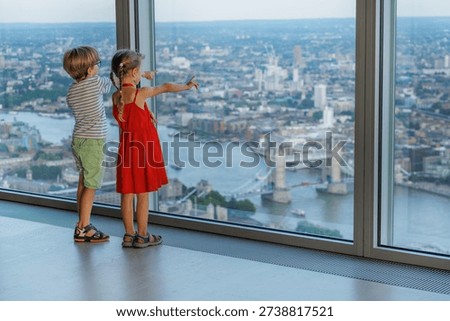Two children point at landmarks while looking out a large window overlooking a cityscape, London, UK