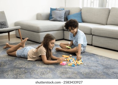Two children settled down on floor in living room surrounded by colorful building blocks, engaged in playful and creative activity, playing favourite game with cubes at home, enjoy their time together