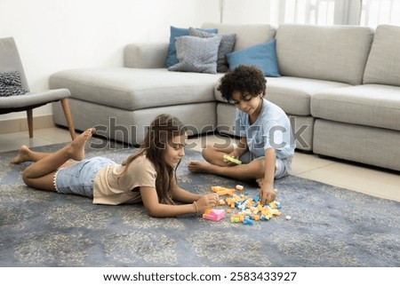 Two children settled down on floor in living room surrounded by colorful building blocks, engaged in playful and creative activity, playing favourite game with cubes at home, enjoy their time together