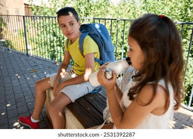 Two children sit on a bench outdoors, one with a backpack and the other drinking from a bottle, enjoying the sunny day. - Powered by Shutterstock