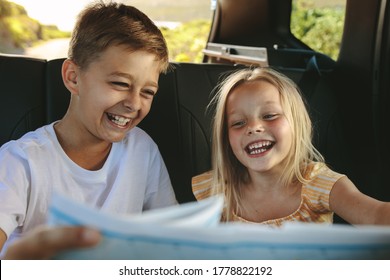 Two Children Sitting On Backseat Of Car Looking At Map And Smiling On Road Trip. Kids Traveling In A Car.