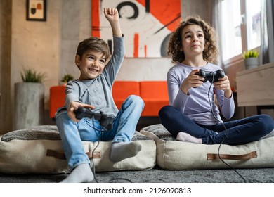 Two children small caucasian brother and sister happy children siblings boy and girl playing video game console using joystick or controller while sitting at home boy is winning - Powered by Shutterstock