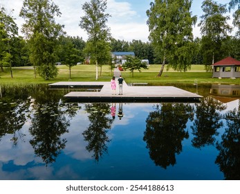 Two children stand on a wooden dock over calm water, reflecting trees and clouds. A red roof house and other buildings are visible among lush greenery. - Powered by Shutterstock