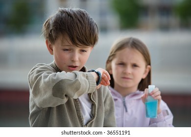 Two Children Stand In An Urban Setting, One Sneezing Into Their Elbow, The Other Holding A Bottle Of Hand Cleanser.