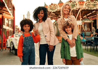 Two children with their mothers came to have fun in the amusement park. An African-American girl and a blond boy with curls are playing and have come to the park to ride a merry-go-round.