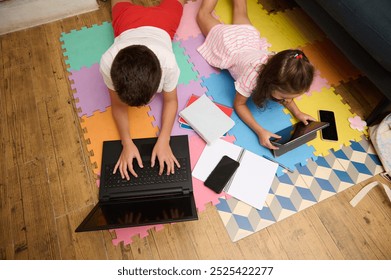 Two children using a laptop and tablet at home, surrounded by colorful play mats, notebooks, and pens, fostering learning and digital interaction. - Powered by Shutterstock