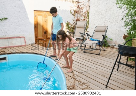 Two children using pool cleaning tools to maintain a backyard swimming pool in daylight.