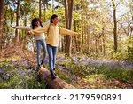 Two Children Walking Through Bluebell Woods In Springtime Balancing On Log