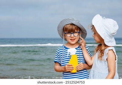 Two children wearing hats and striped outfits are standing by the sea, holding a yellow spray bottle during family vacations - Powered by Shutterstock