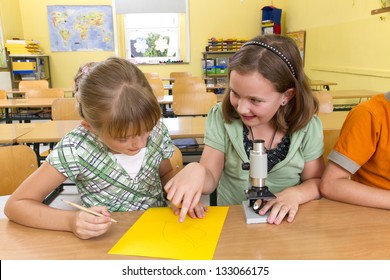 Two Children In A Yellow Classroom. They Are Leaning With A Microscope.