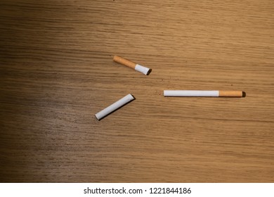 Two Cigarettes On A Wooden Desk Table. One Is Broken, Forming A Triangle Or Arrow Which Could Be A Concept On Smoking Cessation