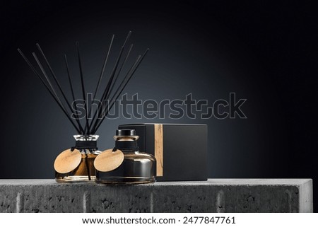 Two clear glass aroma diffuser bottles with amber-colored liquid,  black reed sticks and round blank wooden tags presented on dark elegant backdrop with slender black box and piece of driftwood