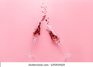 Two clinking champagne glasses with splash of red heart shaped confetti over pink background. Overhead view, copy space. Valentine's Day concept