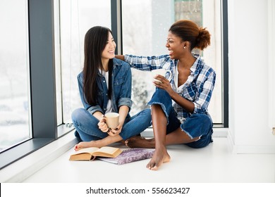 Two Close Friends Of Different Ethnicity Talking And Smiling One To Each Other On The Floor In Small Break Of Studying With Cup Of Coffee In Hands