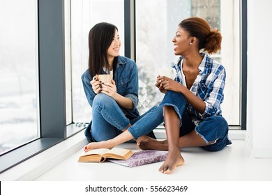 two close friends of different ethnicity talking and smiling one to each other on the floor in small break of studying with cup of coffee in hands