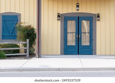 Two closed blue glass doors with white lace curtains and grey trim. There's a window with a blue shutter door. A grey wooden fence with trees, shrubs and flowers. The exterior wall is yellow clapboard - Powered by Shutterstock