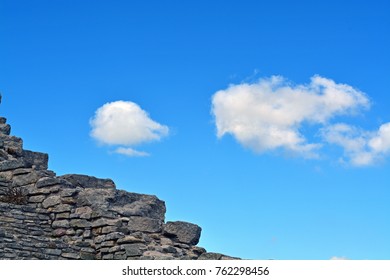 Two clouds floating in a blue sky with a detail of a stone wall in the foreground.