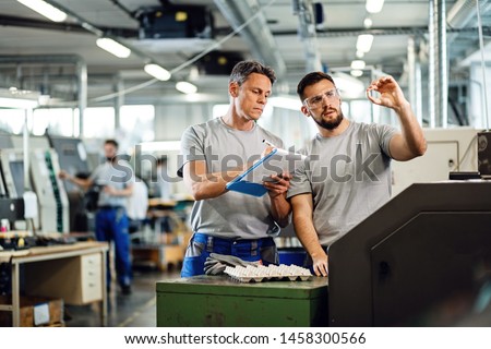 TWO CNC machine operators examining finished products and writing notes in industrial facility. 