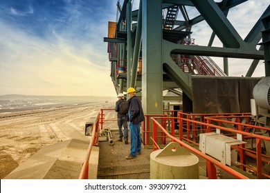 Two Coal Mine Engineers With Protective Helmets Standing On A Huge Drill Machine, Talking And Watching At The Digging Site. Beautiful And Colorful Sky In The Background. Rear View.