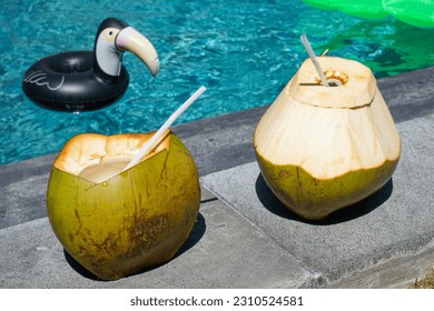 Two coconuts with straws standing in the edge of a pool with a toucan drink float and a green mattress float in the background on a sunny summer day in Bali, Indonesia