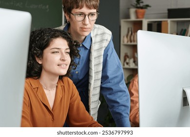 Two colleagues collaborating on computer project in modern classroom setting with bookshelves and large monitors, one person standing and the other seated