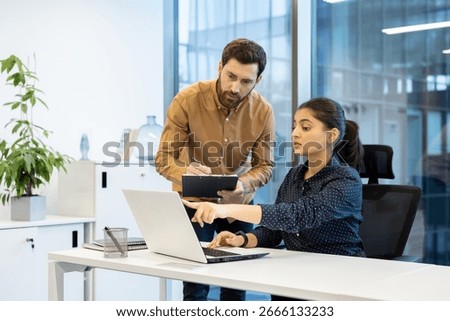 Two colleagues discuss a project while working on a laptop in a professional setting. The interaction showcases teamwork and productivity in a modern office environment.