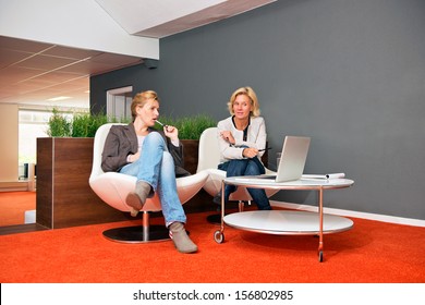 Two Colleagues Discussing A Project In An Informal Office Setting, With Laptop, Notes And Both Women Casually Dressed